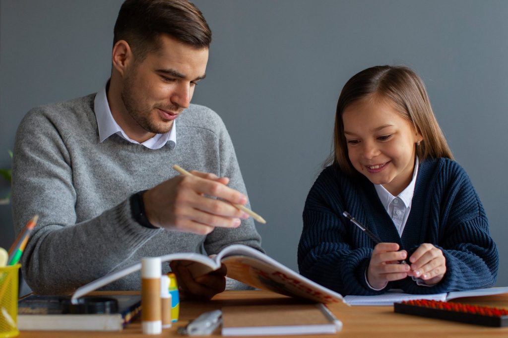 Un tuteur adulte montre quelque chose du doigt dans un livre ouvert à une jeune élève souriante, assise côte à côte à un bureau avec des fournitures scolaires.